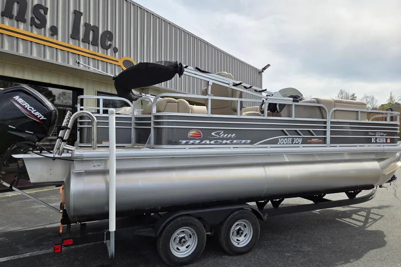 Slide: The Image of 2023 Sun Tracker Fishin' Barge 20 DLX pontoon boat on trailer, parked outside a building. - 2
