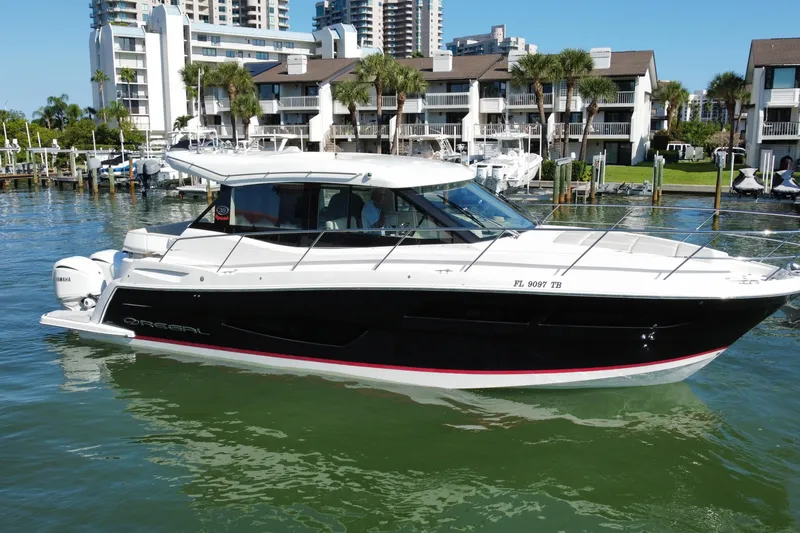 The Image of 2022 Regal 38 XO boat docked in marina, surrounded by modern buildings and palm trees. - 0
