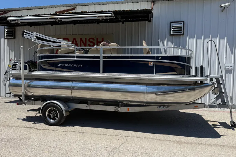 Slide: The Image of 2023 Starcraft LX 18 F pontoon boat on trailer, parked outside a metal building. - 2