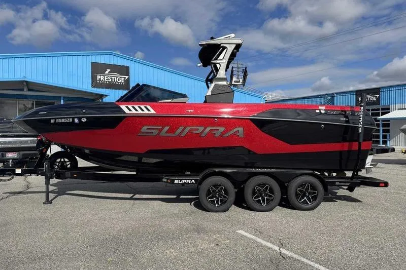 The Image of 2022 Supra SE boat in red and black on a trailer, parked outside a blue building. - 1