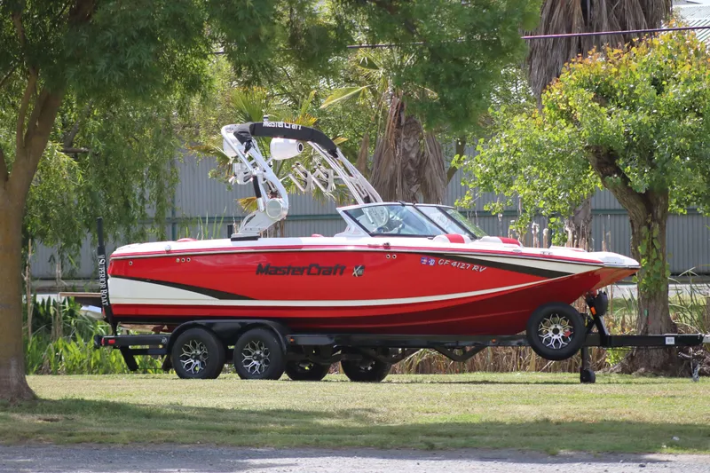 The Image of 2014 MasterCraft X-25 boat on trailer, parked outdoors among trees. - 0