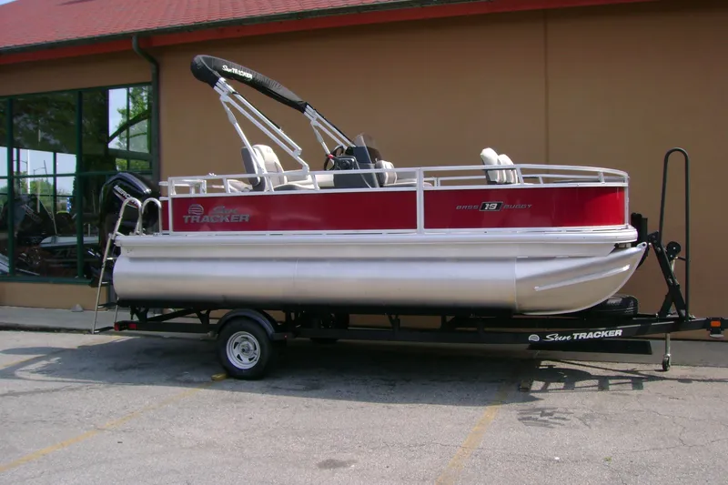 Slide: The Image of 2026 Sun Tracker Bass Buggy 18 DLX pontoon boat on trailer, parked outside a building. - 2