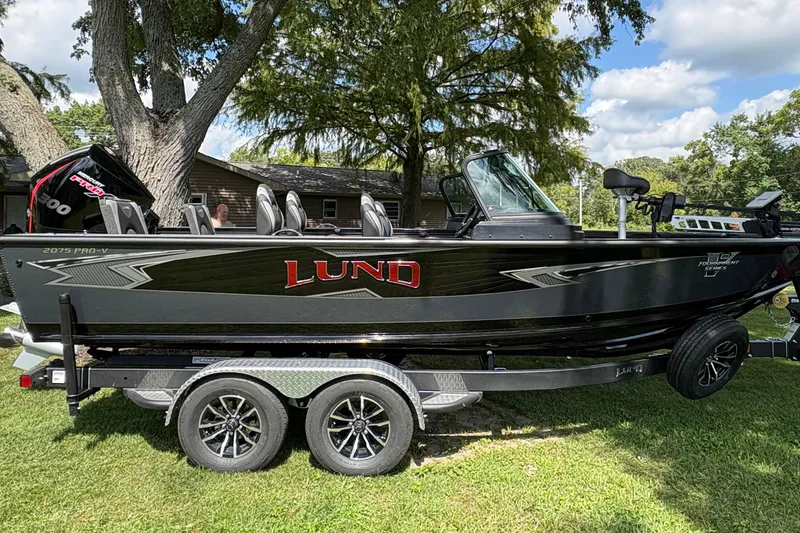 The Image of 2026 Lund 2075 Pro-V Sport boat on trailer, parked on grass under trees. - 1
