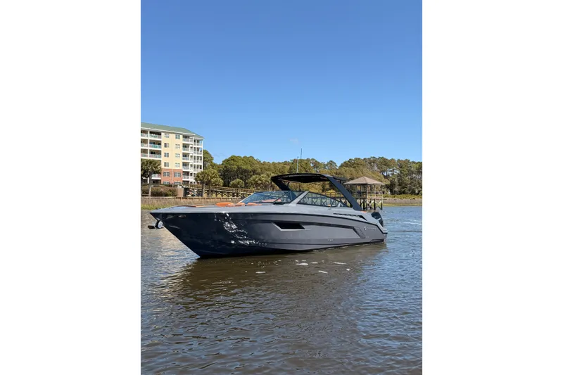 Slide: The Image of 2021 Cruisers Yachts 338 Bow Rider on calm water, with buildings and trees in the background. - 2