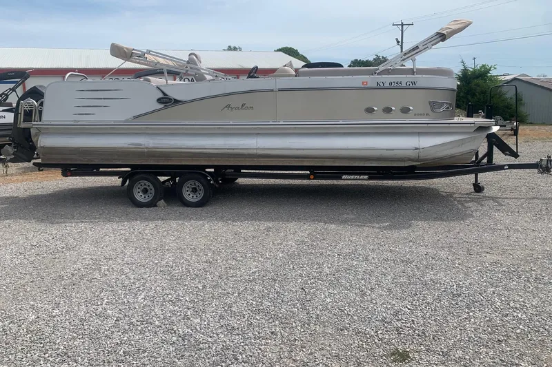 The Image of 2015 Avalon Windjammer Elite 25' pontoon boat on trailer, parked on gravel. - 0