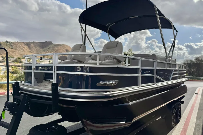 Slide: The Image of 2024 Ranger Reata 223F pontoon boat with canopy, parked on a trailer, scenic background. - 3