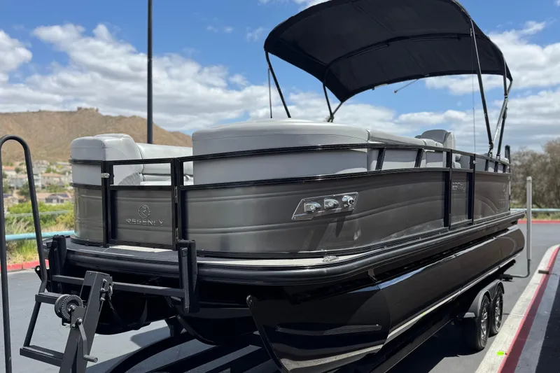 Slide: The Image of 2024 Regency 230 DL3 pontoon boat with canopy, parked on a trailer, scenic background. - 3