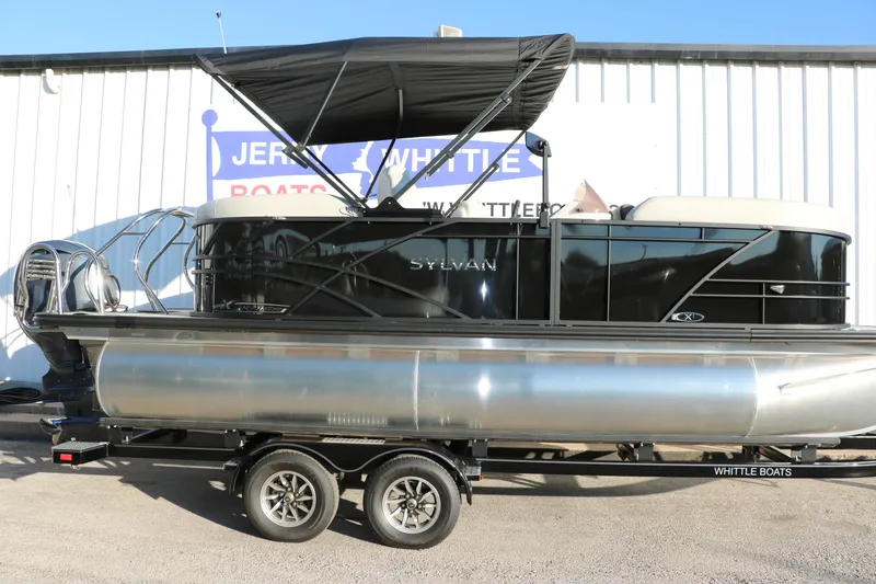 Slide: The Image of 2025 Sylvan Mirage X1 pontoon boat with black canopy on trailer, parked outside dealership. - 9