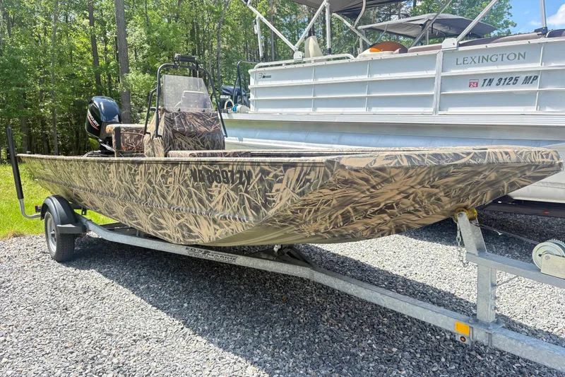 The Image of 2022 Lowe Roughneck 1860CC camo boat on trailer, parked outdoors near trees. - 1