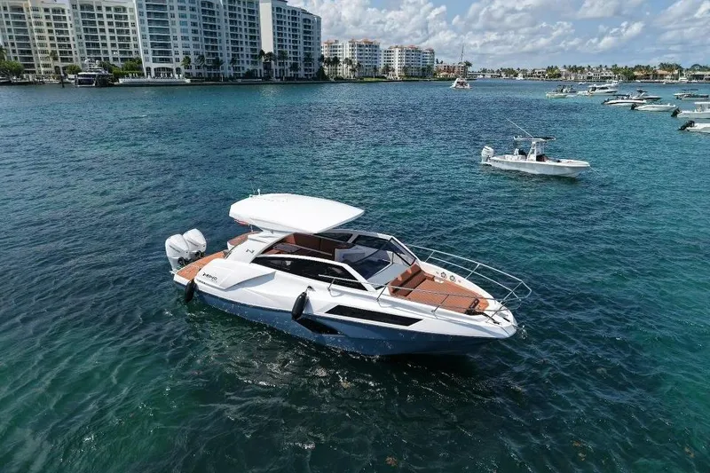 Slide: The Image of 2025 NX Boats 340 Sport Coupe cruising on clear blue water near coastal buildings. - 12