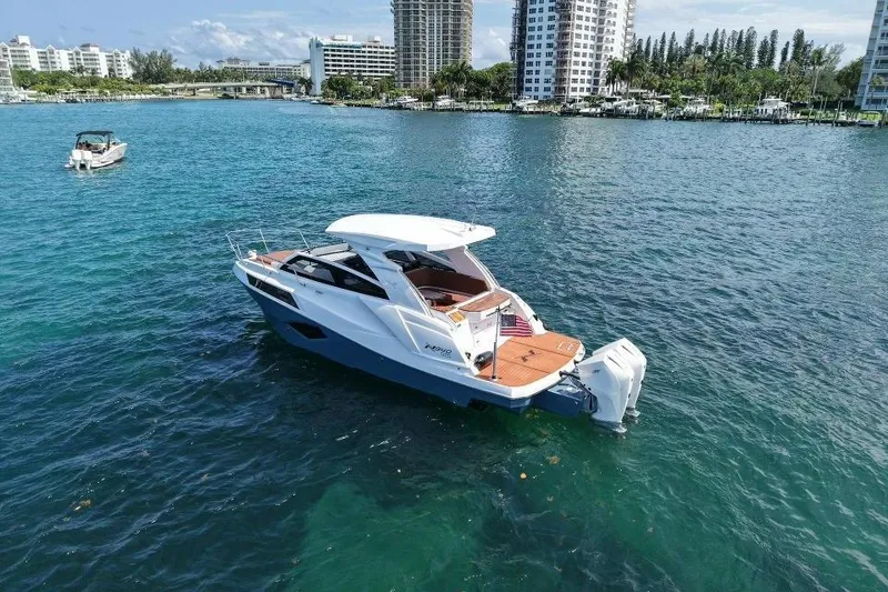 Slide: The Image of 2025 NX Boats 340 Sport Coupe cruising on clear blue water near city skyline. - 9