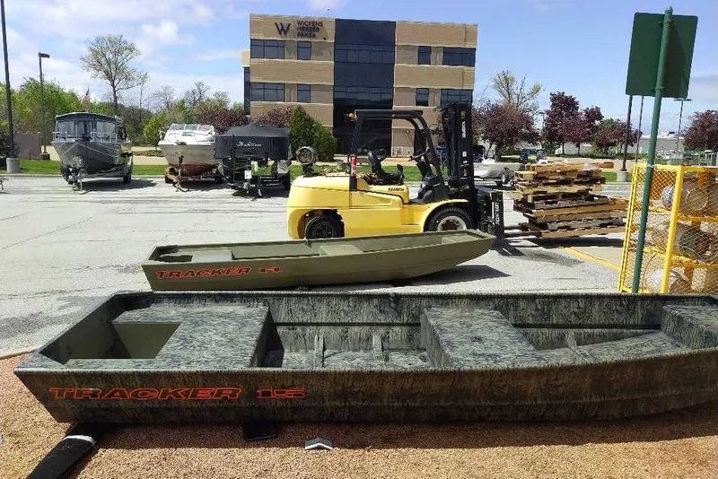 The Image of 2026 Tracker Grizzly 15 Jon boat displayed outdoors near forklift and other boats. - 0