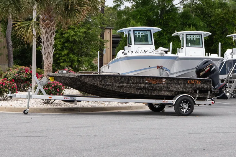 The Image of 2026 Excel 203 Bay Pro Elite boat on trailer, parked near palm trees and flowers. - 1
