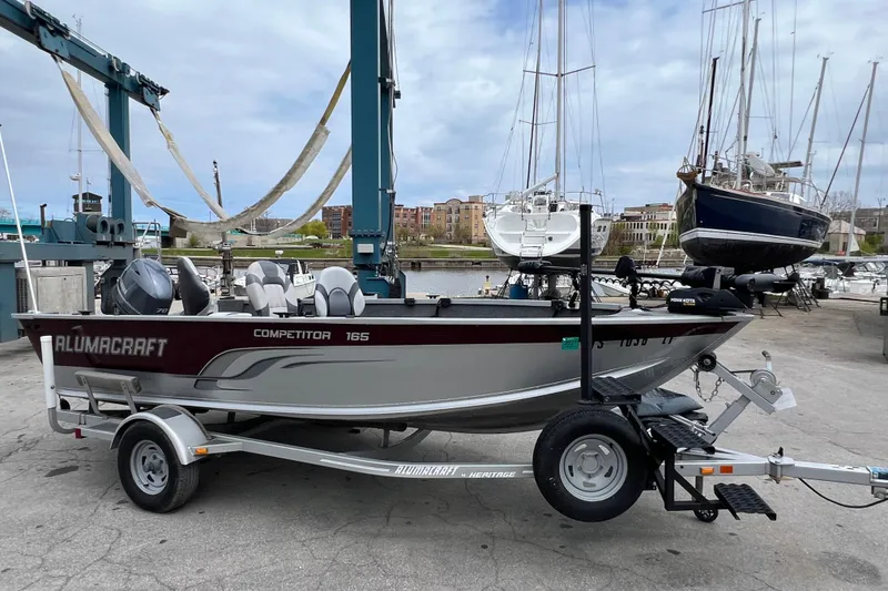 Slide: The Image of 2014 Alumacraft Competitor 165 Tiller boat on trailer at marina, surrounded by sailboats. - 3