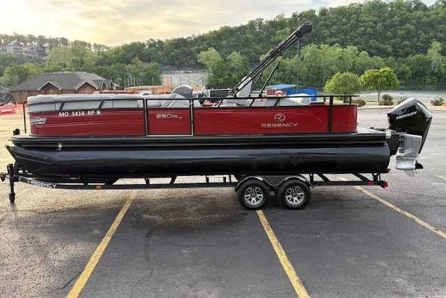 Slide: The Image of 2024 Regency 250 DL3 pontoon boat on trailer in parking lot, surrounded by trees. - 8