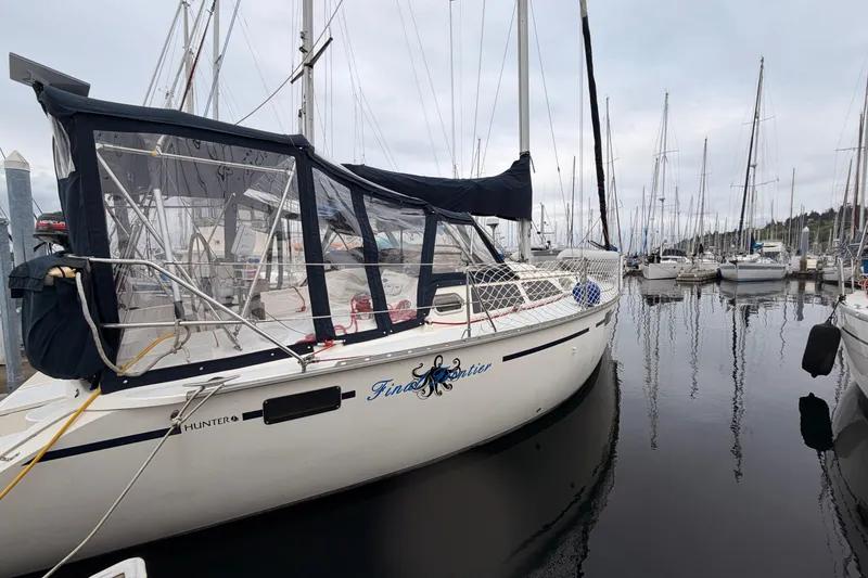 The Image of 1990 Hunter 37.5 sailboat docked in a marina, featuring a blue canopy and calm waters. - 0