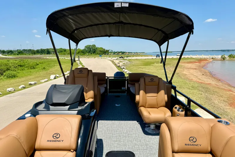 Slide: The Image of 2026 Regency 230 DL pontoon boat with tan seating, docked by a scenic lakeside. - 13