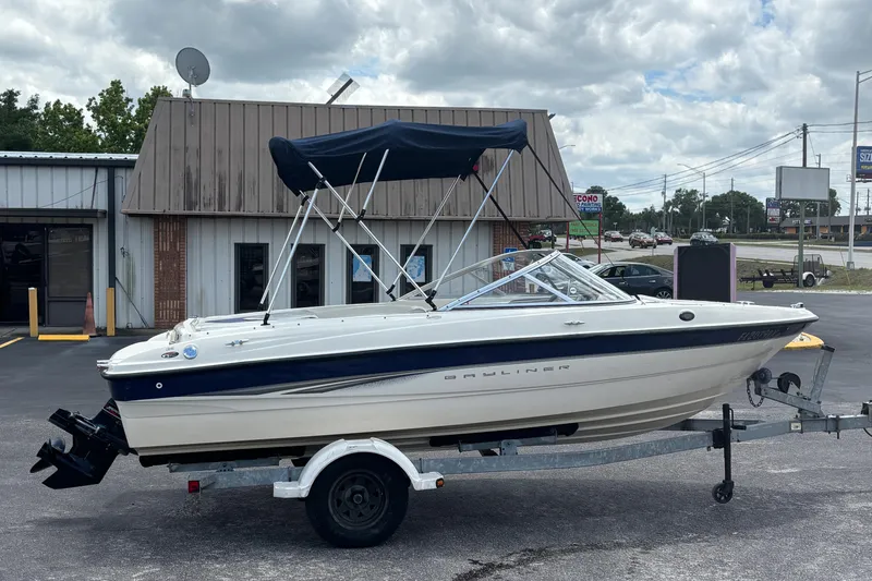 Slide: The Image of 2003 Bayliner 185 Capri boat on trailer, parked outside a building under cloudy skies. - 6