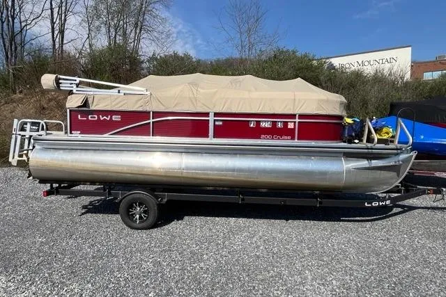The Image of 2023 Lowe Ultra 200 Cruise pontoon boat on trailer, covered and parked outdoors. - 0