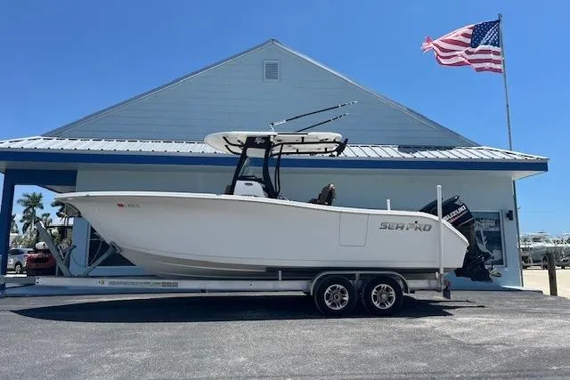 The Image of 2019 Sea Pro 259 Center Console boat on trailer, parked by a building with an American flag. - 1