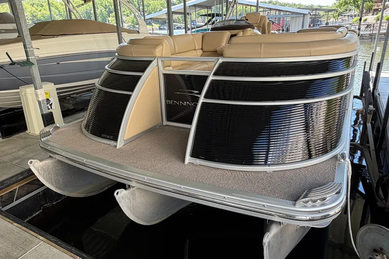 Slide: The Image of 2011 Bennington 2575QCW OB pontoon boat docked, featuring beige seating and sleek black paneling. - 4