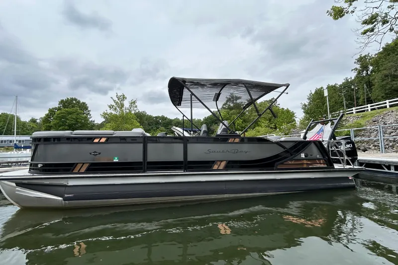Slide: The Image of 2025 South Bay 525UL393 pontoon boat docked on a calm lake under cloudy skies. - 11