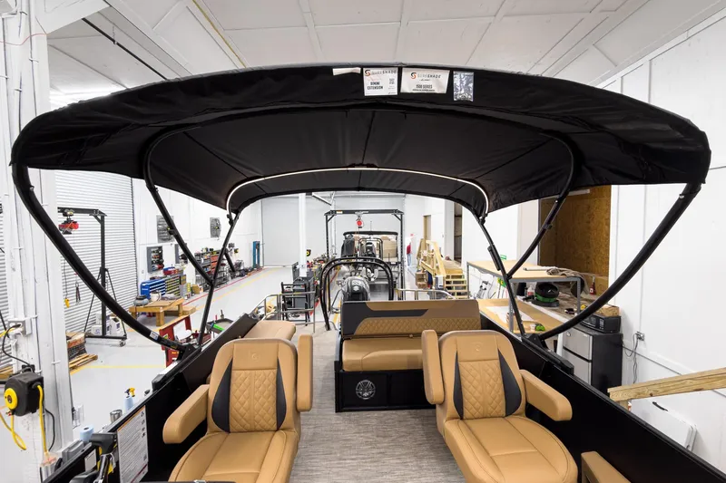 Slide: The Image of 2026 Godfrey Sweetwater 22' Tritoon with tan seating and black canopy in a workshop. - 25