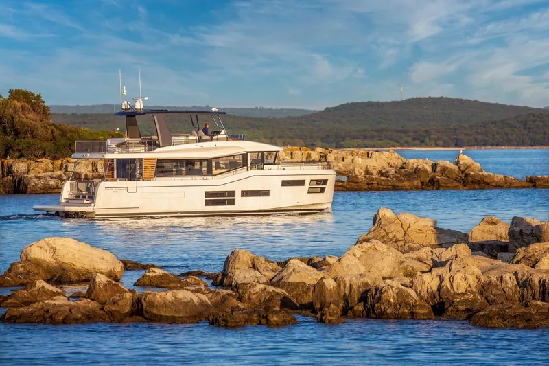 The Image of 2022 Beneteau Grand Trawler 62 cruising near rocky shoreline under blue sky. - 0