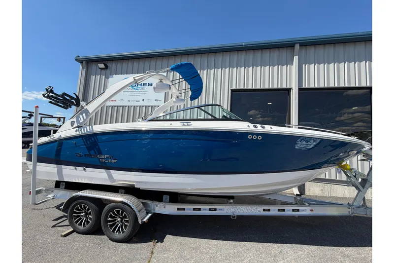 Slide: The Image of 2026 Regal LS6 Surf boat on trailer, blue and white design, parked outside a building. - 1