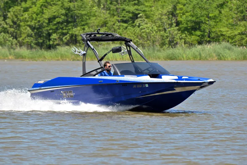 The Image of Blue 2014 Axis A22 boat cruising on a lake with lush green trees in the background. - 1
