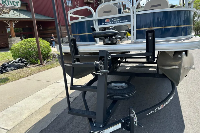 Slide: The Image of 2024 Sun Tracker Bass Buggy 16 XL Select pontoon boat on trailer, parked near a shop. - 12