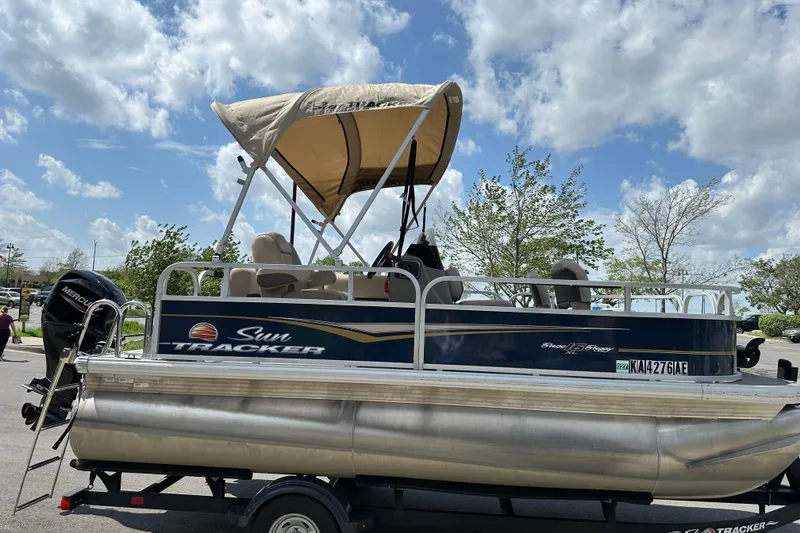 Slide: The Image of 2024 Sun Tracker Bass Buggy 16 XL Select pontoon boat with canopy, parked outdoors. - 2