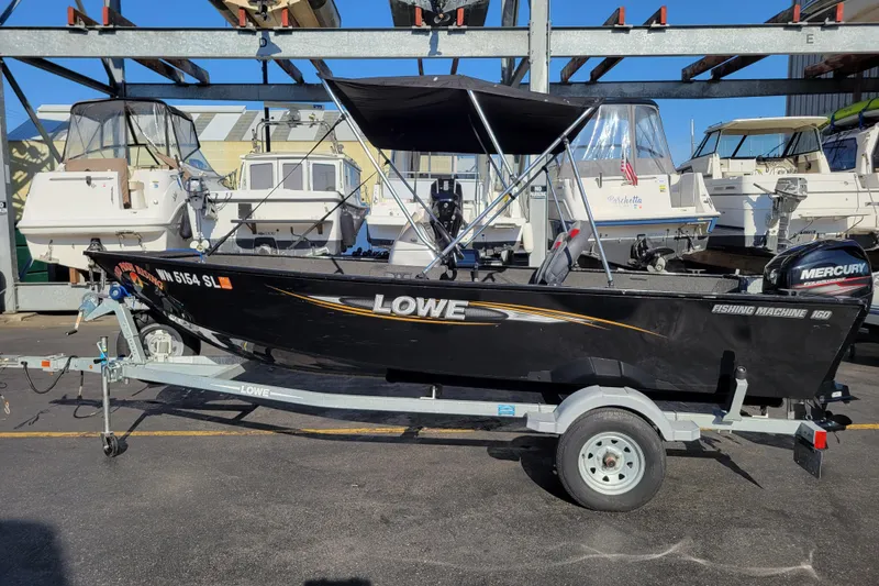 Slide: The Image of 2017 Lowe Fishing Machine 160S boat on trailer, parked in marina with other boats. - 2