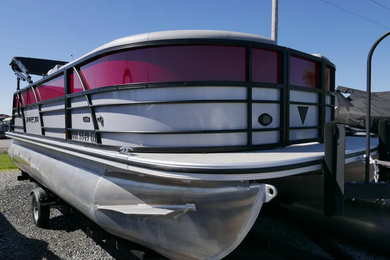 Slide: The Image of 2023 Trifecta C Series pontoon boat with red accents, parked on gravel. - 2