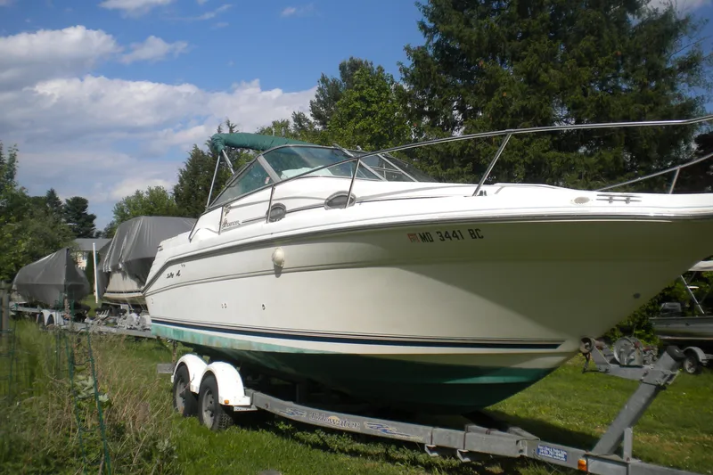 The Image of 1996 Sea Ray 270 Sundancer boat on trailer, surrounded by greenery and trees. - 0