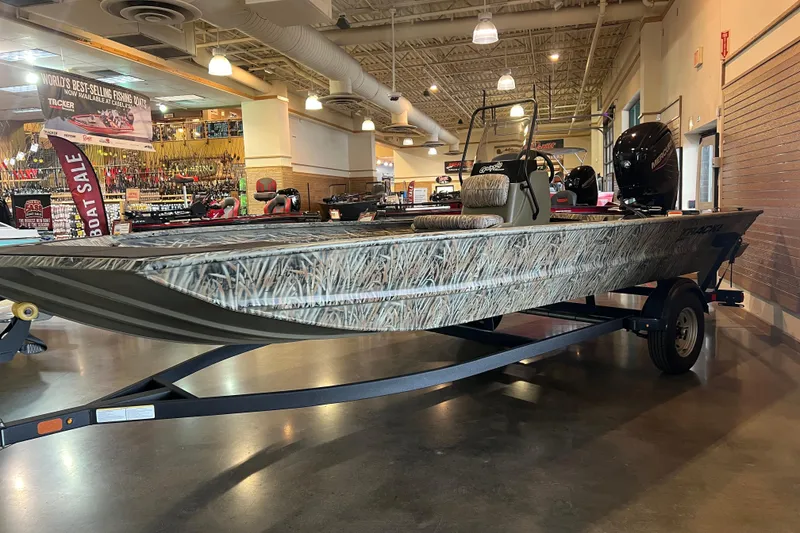 The Image of 2026 Tracker Grizzly 1860 CC boat displayed indoors, featuring camouflage design and Mercury outboard motor. - 0