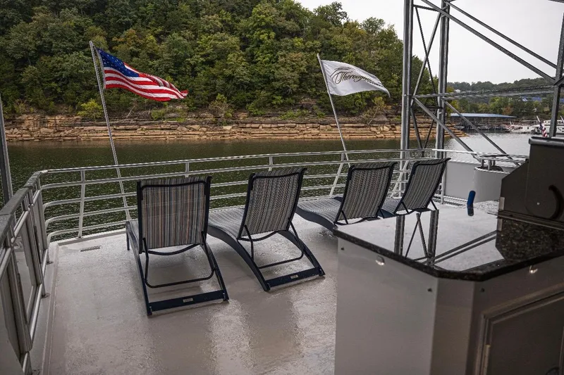 Slide: The Image of Deck of 2020 Thoroughbred 20 x 102 houseboat with lounge chairs and flags, overlooking a scenic river. - 56