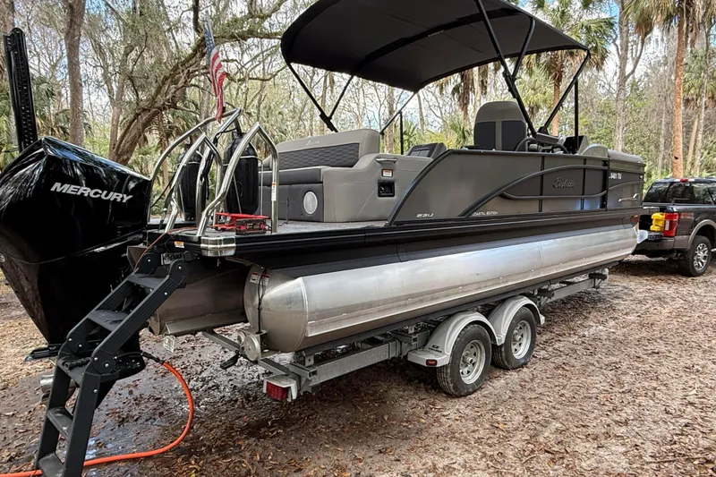 Slide: The Image of 2023 Barletta Corsa 23U pontoon boat on trailer in wooded area. - 2