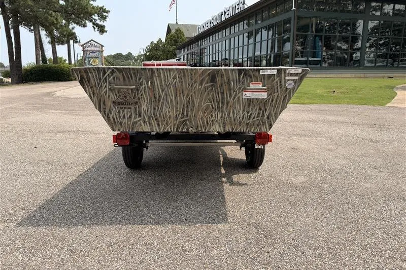 Slide: The Image of 2026 Tracker Grizzly 15 Jon boat on trailer, parked outside a boat center. - 4