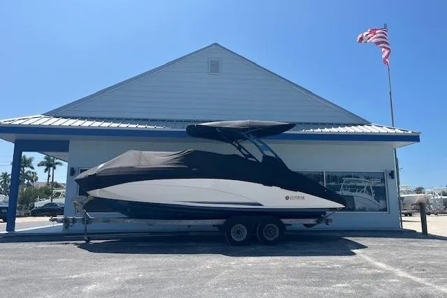 Slide: The Image of 2022 Yamaha 242SE boat covered, parked on trailer outside building with American flag. - 2