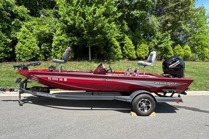 The Image of 2024 Ranger RT188 boat in red on a trailer, parked on a road with greenery background. - 0