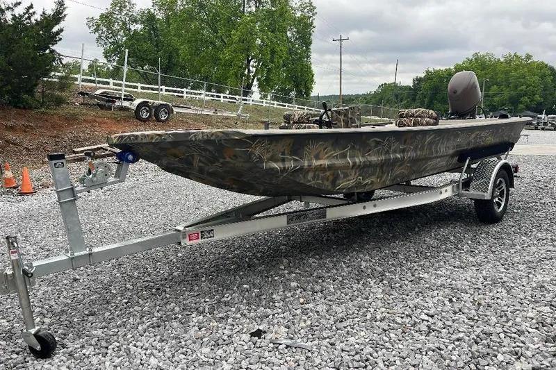 Slide: The Image of 2026 War Eagle 754VS Stick Steer boat on trailer, camo design, parked on gravel. - 2