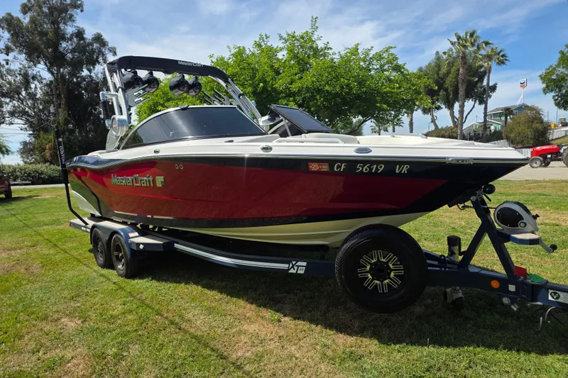 Slide: The Image of 2019 MasterCraft XT23 boat on trailer, parked on grass with trees in background. - 1