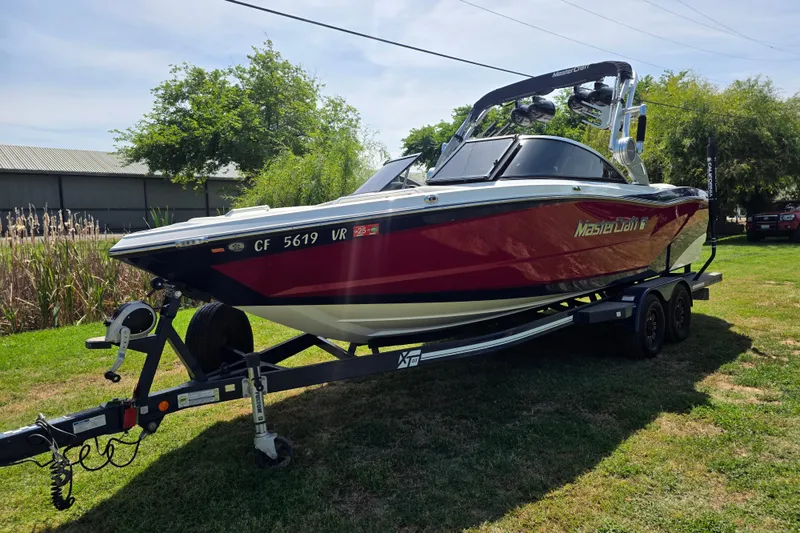 Slide: The Image of 2019 MasterCraft XT23 boat on trailer, parked on grass, with trees in the background. - 2
