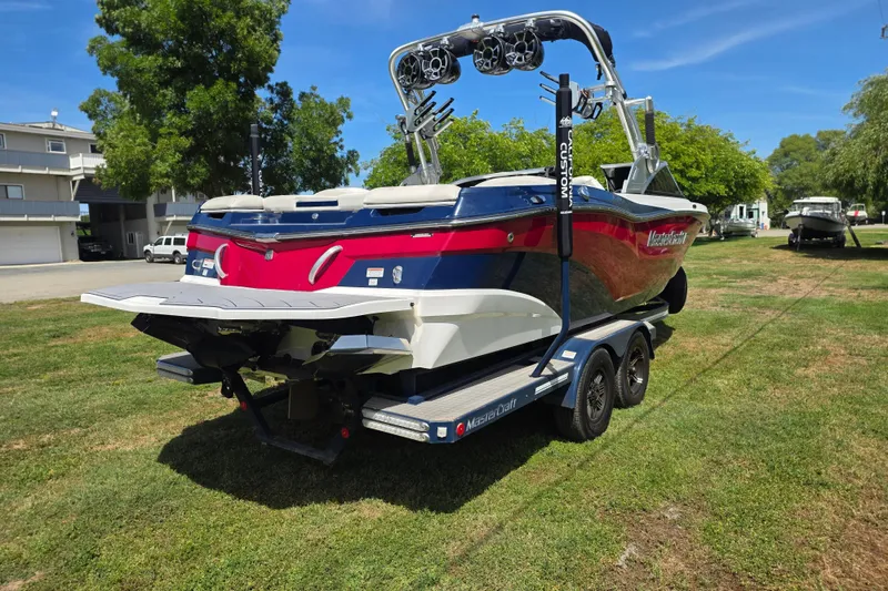 Slide: The Image of 2019 MasterCraft XT23 boat on trailer, parked on grass with trees and buildings nearby. - 6