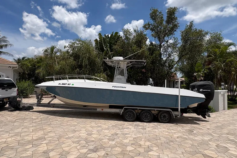 The Image of 2001 Fountain 31 Center Console boat on trailer, parked in a driveway with lush greenery. - 0