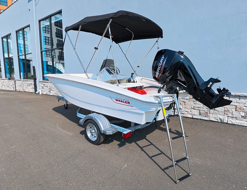 Slide: The Image of A 2027 Boston Whaler 130 Super Sport on a trailer. It features a black Bimini top and a Mercury outboard motor. - 3