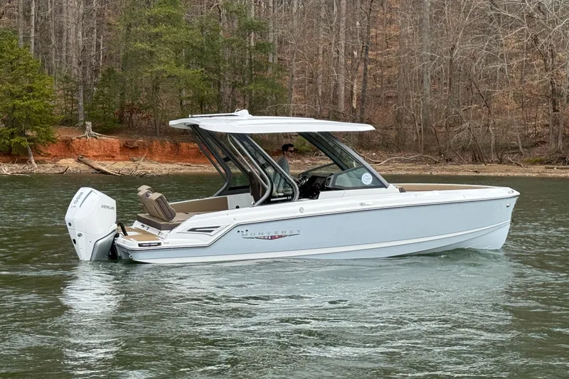 Slide: The Image of 2026 Monterey Elite 25 OB boat cruising on a serene lake with wooded shoreline. - 2