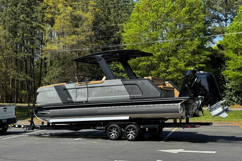 Slide: The Image of 2026 Revel RS25L pontoon boat on trailer, parked outdoors with lush green trees in background. - 19