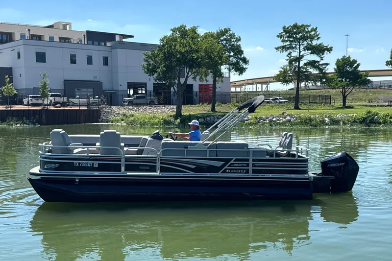 The Image of 2022 Ranger 223FC pontoon boat on a calm lake near a modern building. - 1